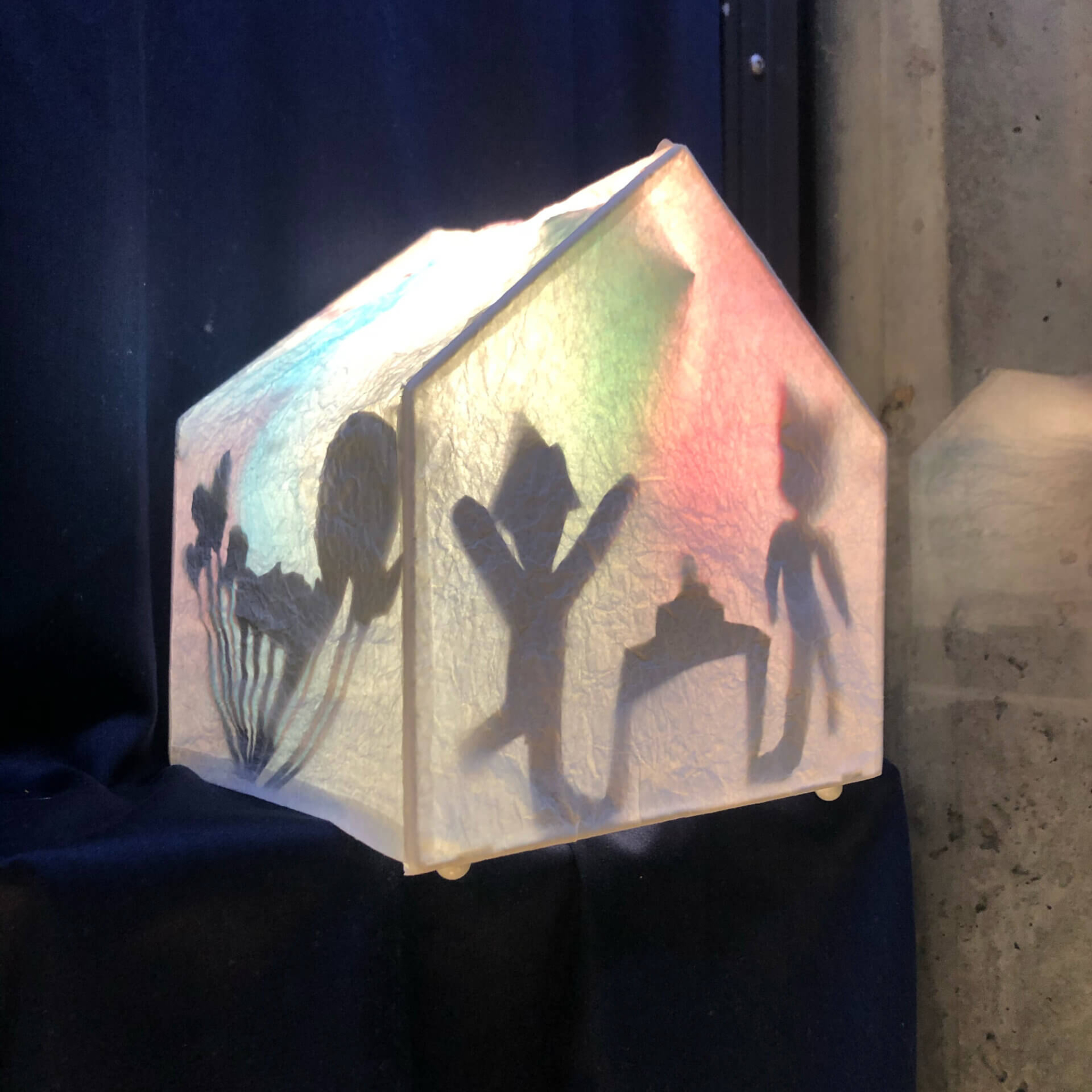 Sisältä valaistu paperinen talonmuotoinen lyhty, jossa näkyy varjokuvia ihmishahmoista.