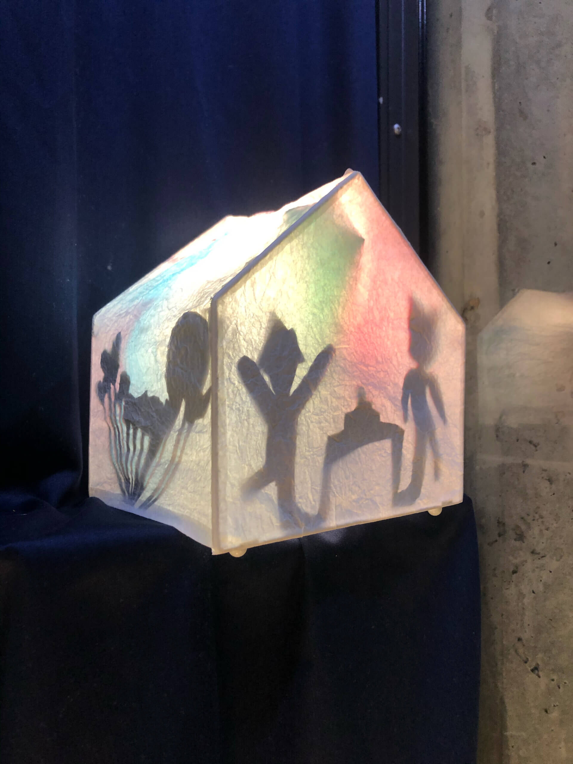 Sisältä valaistu paperinen talonmuotoinen lyhty, jossa näkyy varjokuvia ihmishahmoista.
