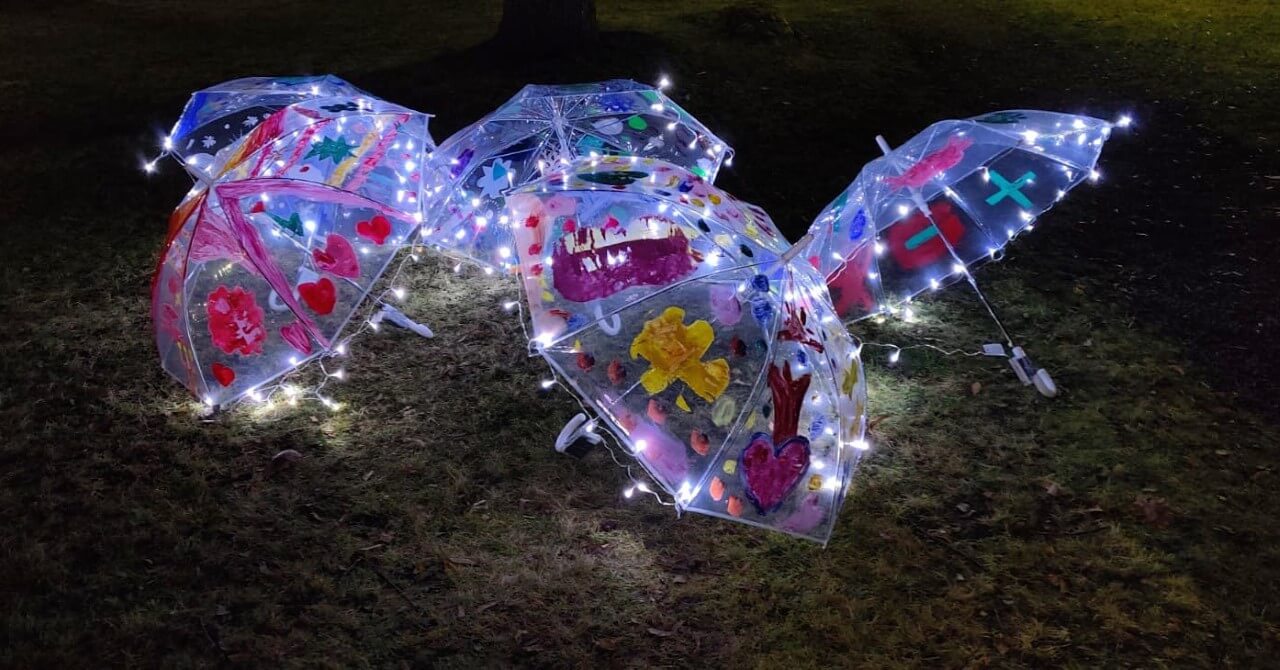 Valonauhalla valaistuja lasten maalaamia läpinäkyvästä muovista tehtyjä sateenvarjoja nurmikolla.
