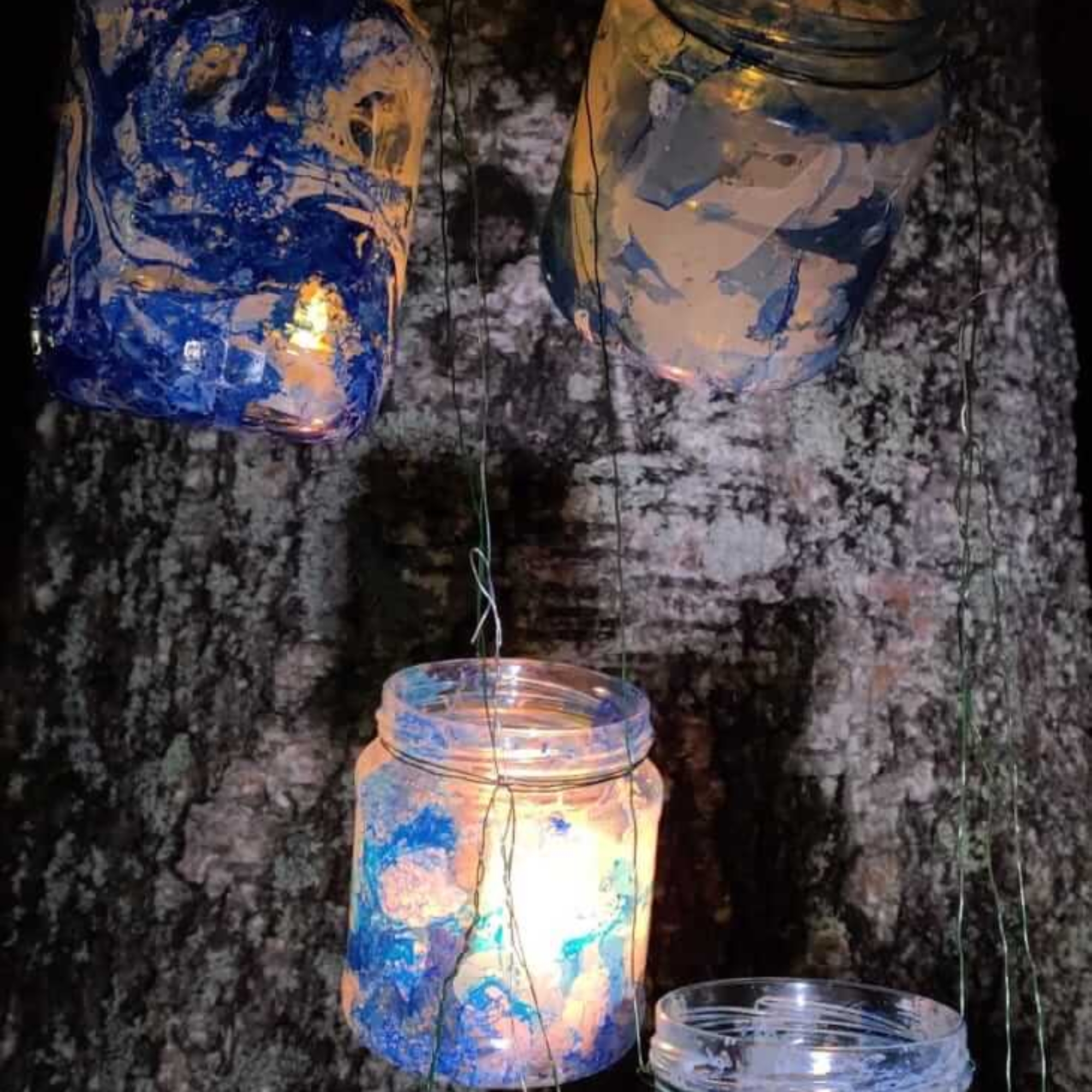 Lasten maalaamia lasipurkeista tehtyjä kynttilälyhtyjä ripustettuna puun runkoon.