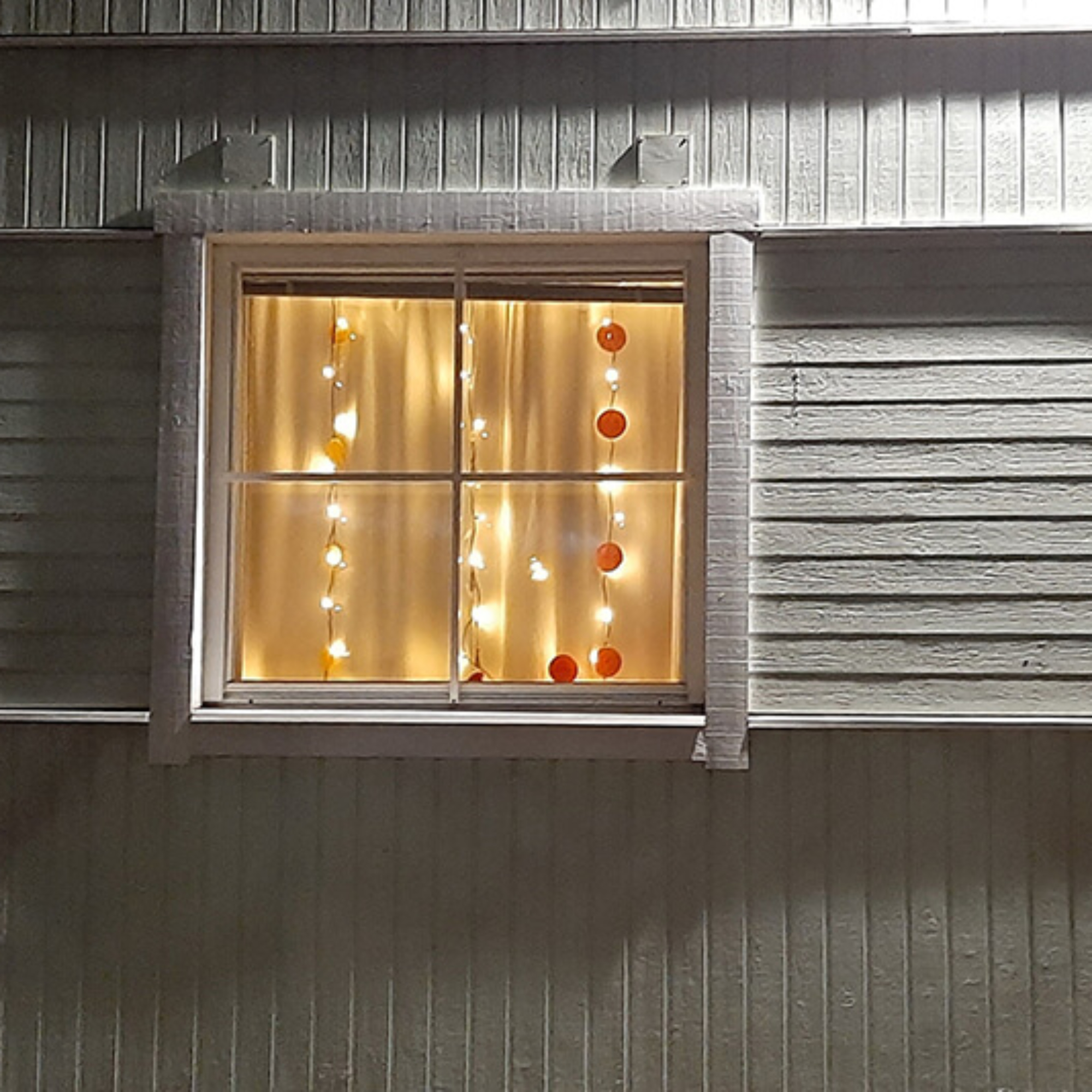 Erilaisia lasten tekemiä valonauhoja ja valaistuja lyhtyä vaalean puutalon kolmessa ikkunassa.