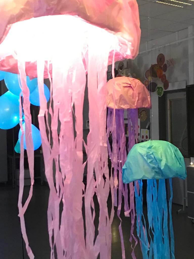 Pastellisävyisistä silkkipapereista tehtyjä ja valaistuja meduusoja roikkumassa luokkahuoneen katosta.