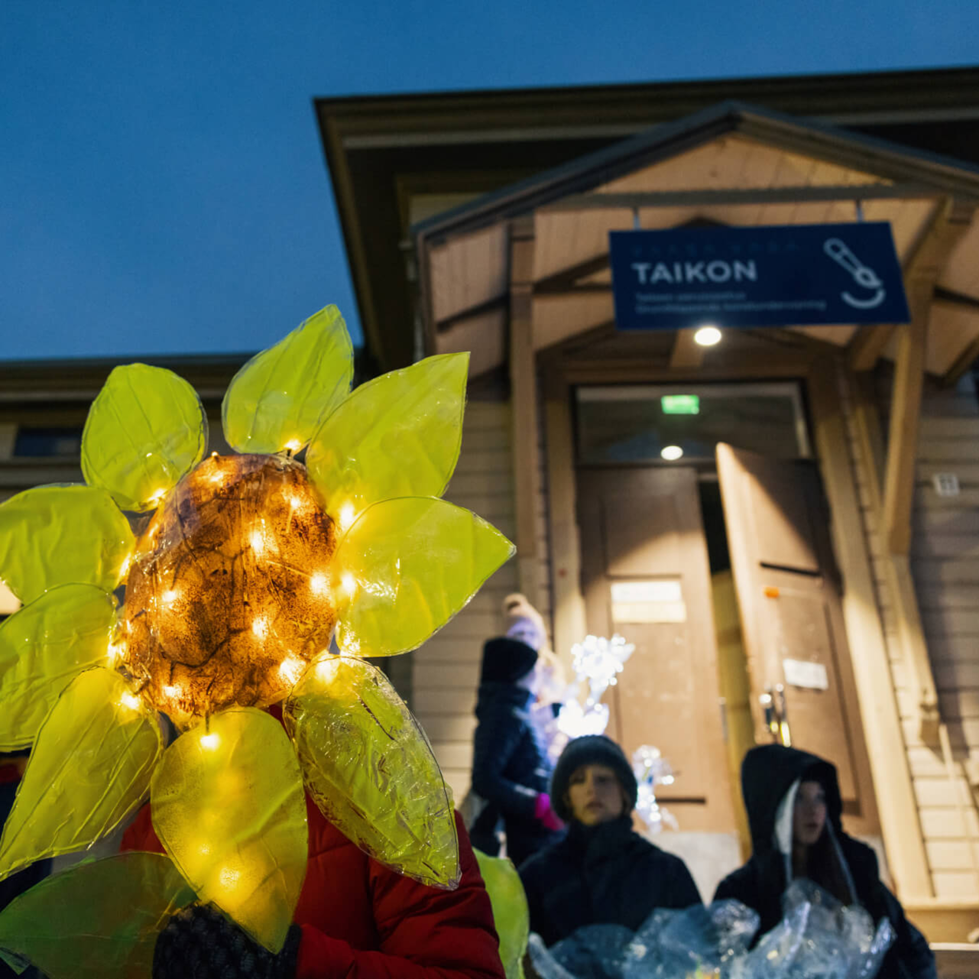 Valonauhalla valaistu keltainen kukkateos ja lapsia Vaasan TAiKonin puutalon edustalla.
