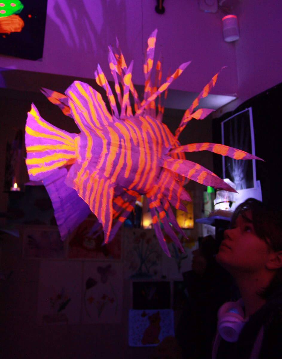 Uv-valolla valaistu violetti-oranssi-raitainen kala-taideteos roikkumassa katosta.