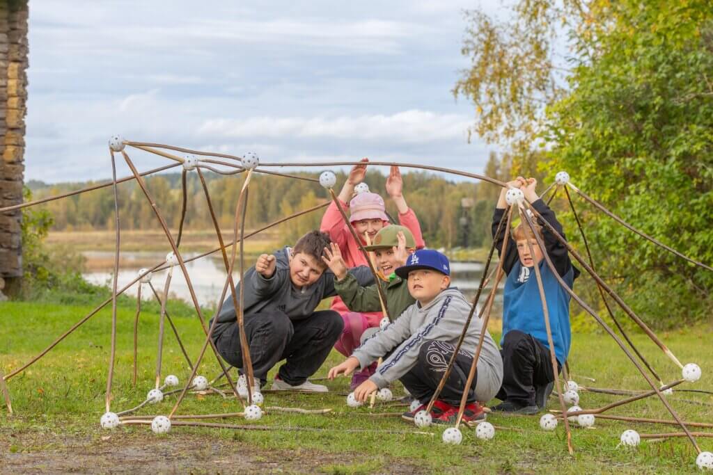 Viisi lasta kyykyssä rakentamansa pajurakennelman sisällä syksyisessä järvimaisemassa.