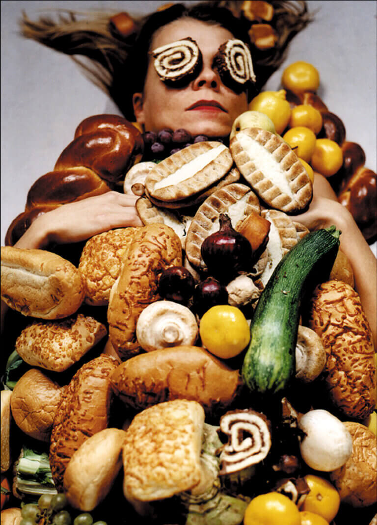 VAlokuvateos, jossa näkyy naisen kasvot vihannesten ja juuresten alta.