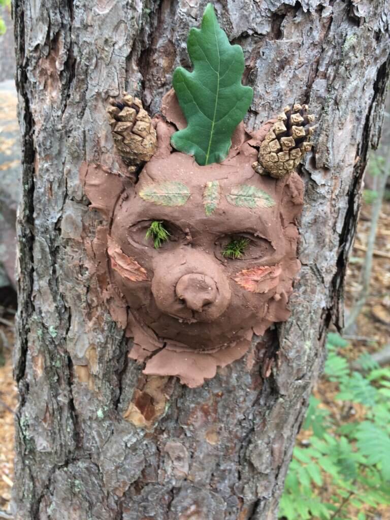 Männyn runkoon savesta tai vastaavasta massasta tehty karhun päätä esittävä kolmiulotteinen teos, jolla on kävyt korvina ja tammenlehti päässä.